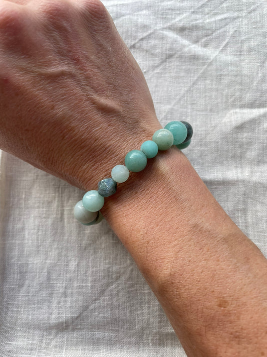 A close-up of a person's wrist wearing a beaded stretch bracelet made of light aqua and white polished stones, resting on a textured white linen fabric background.